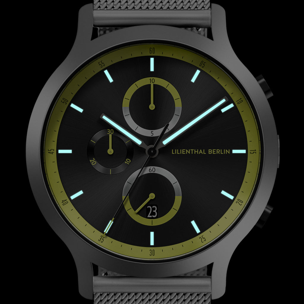 Modern black chronograph watch with green accent markers, mesh strap, and luminous hands