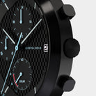 Close-up of a black chronograph watch with a carbon fiber dial, subdials, and date display.