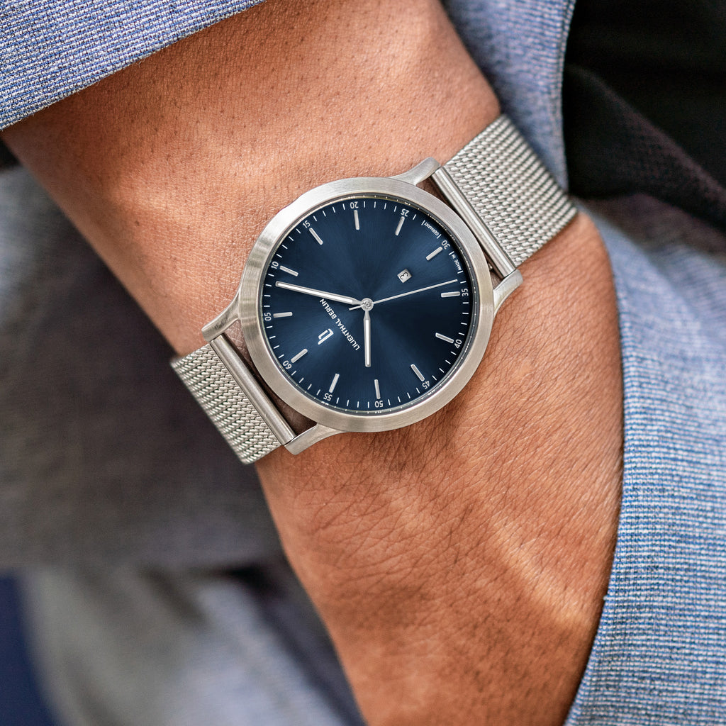 Minimalist silver mesh wristwatch with dark blue dial on wrist, worn with light blue suit