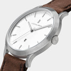 Minimalist silver wristwatch with white dial, brown leather strap, and date display