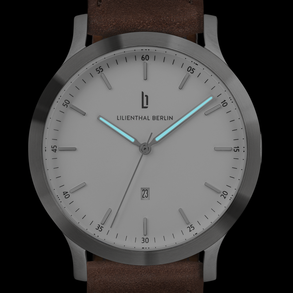 Minimalist silver watch with white dial, glowing blue hands, date display, brown leather strap