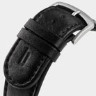 Close-up of a black leather watch strap with a silver buckle on a light background