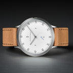 Minimalist silver watch with white dial and tan leather strap on dark textured surface