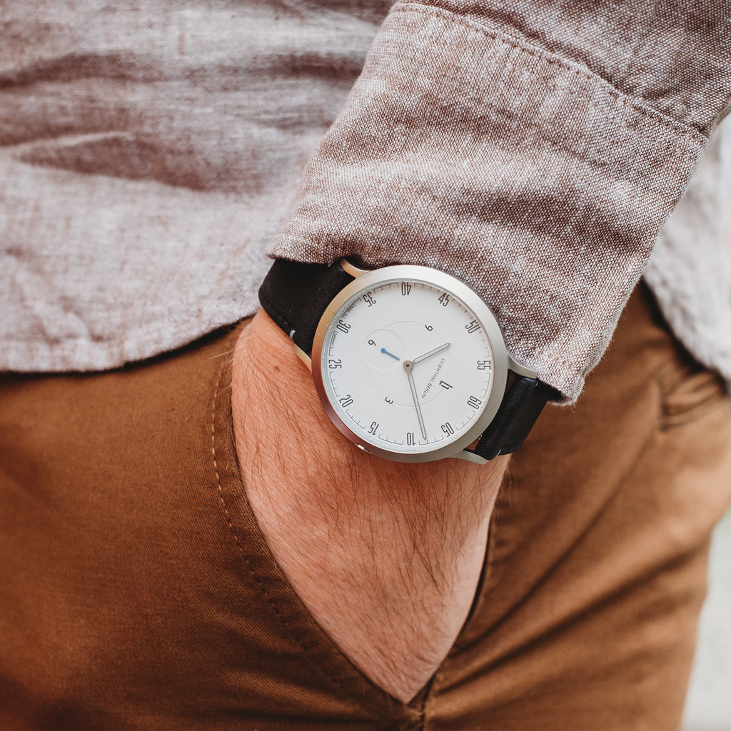 Minimalist wristwatch with white dial and black strap on wrist, casual outfit background