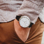 Minimalist wristwatch with white dial and black strap on wrist, casual outfit background