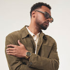 Man in olive jacket wearing square sunglasses and gold wristwatch against neutral background